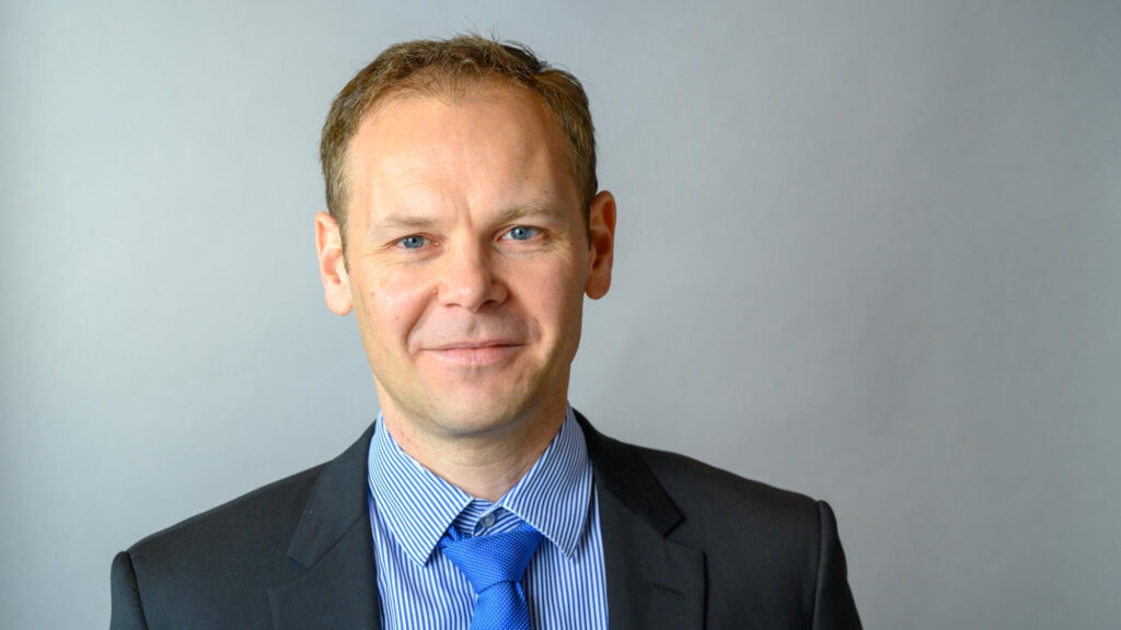 Das Foto zeigt Christoph Müller. Er hat blonde, kurze Haare und blaue Augen. Er trägt ein dunkelgraues Sakko und darunter ein weiß-blau liniertes Hemd mit einer weiß-blau gepunkteten Krawatte.