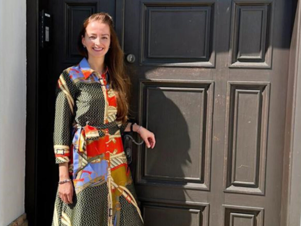 Das Bild zeigt Julia Rainert. Sie hat braune, brustlange Haare und trägt ein buntes Kleid in den Farben rot, blau und mit grün-gelben Linien. Sie steht an einer Haustür und lächelt in die Kamera.