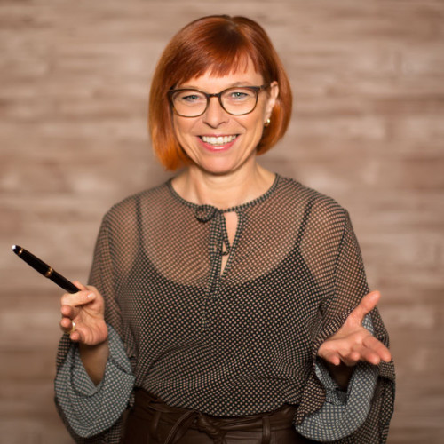 Das Bild zeigt Astrid Leutner, eine Trainerin aus dem ZamStarten Gründerzentrum. Sie hat rot gefärbte halslange Haare, eine schwarze Brille und lächelt. Sie streckt ihre Hände öffnend dem Betrachtenden entgegen.