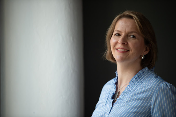 Auf dem Foto ist eine Frau mit halslangen braunen, glatten Haaren zu sehen. Sie trägt Kreolen-Ohrringe und eine hellblaue Bluse mit weißen Streifen. Sie lächelt in die Kamera.