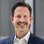 Das Bild zeigt den ZamStarten Trainer Klaus Obermaier. Er hat relativ kurze schwarz-graumelierte Haare und einen gepflegten Bart. Er trägt ein blaues Nadelstreifensakko mit weißem Hemd darunter.