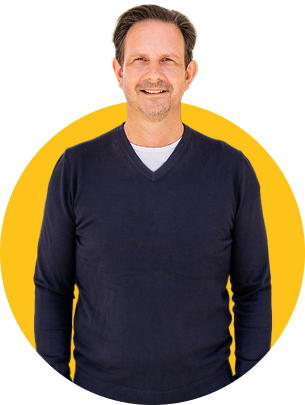 Das Bild zeigt den ZamStarten Trainer Klaus Obermaier. Er hat kurze, leicht abstehende Haare mit grauen Ansätzen, trägt einen dunkelblauen Pullover und darunter ein weißes Hemd.