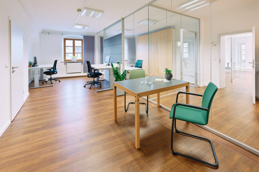 Ein Büroraum mit 3 Schreibtischen in weiß und grün, davor Bürostühle in Schwarz und dazwischen Pflanzen in großen Behältnissen.