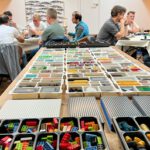 Zwei Gruppen von Männern sitzen an zwei Tischen und arbeiten mit dem Konzept LEGO® Serious Play®, also mithilfe von Lego-Bausteinen Klarheit im Denken und Planen zu schaffen. Im Vordergrund ist ein Tisch mit vielen verschiedenen Lego-Bausteinen zu sehen.